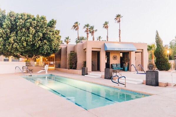 A pool area with a spa and therapy building, surrounded by palm trees and a large tree.
