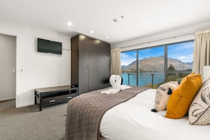 A bedroom with a large bed, a TV mounted on the wall, and a view of mountains and water through a large window.
