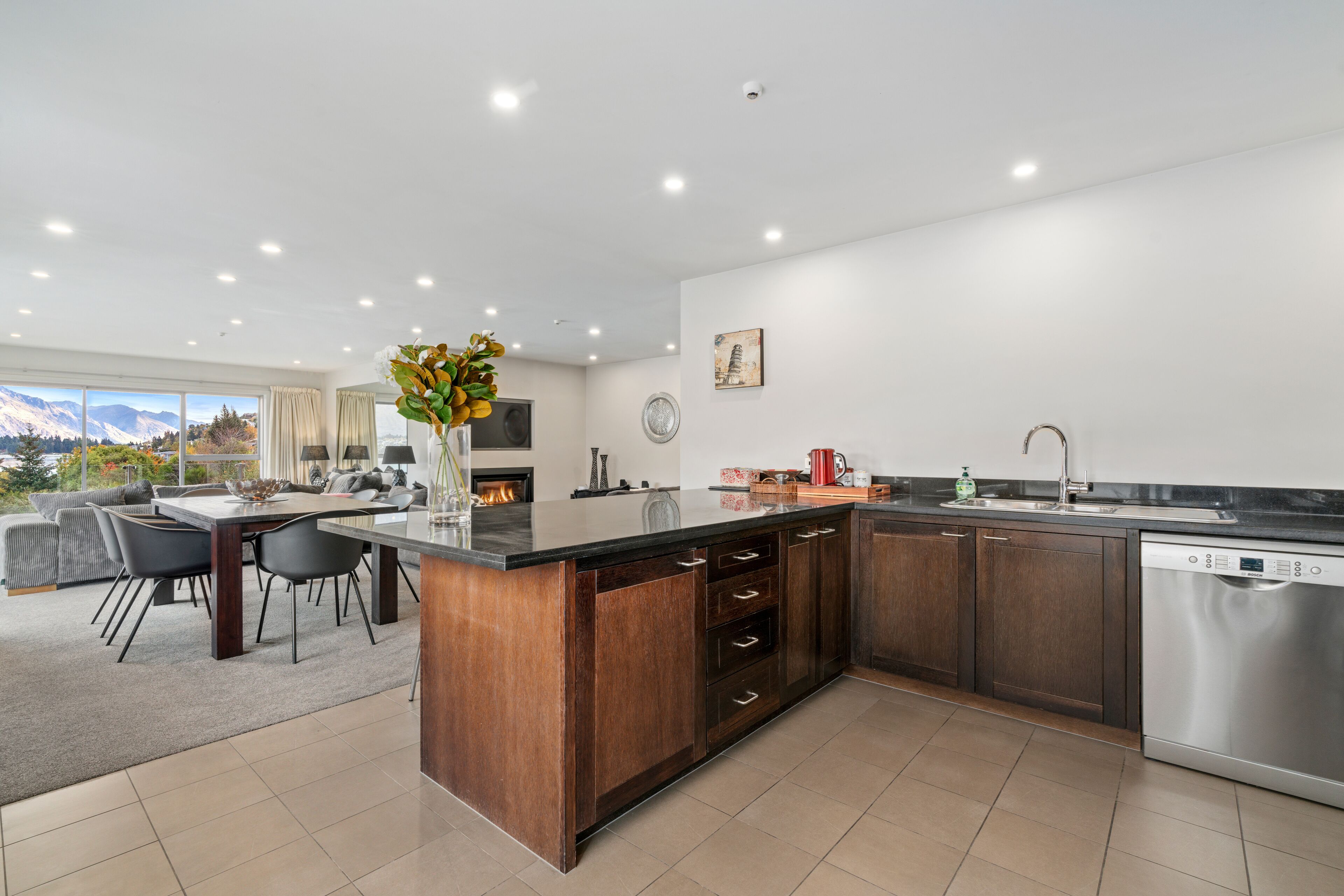 A modern kitchen with a central island, dark wooden cabinets, stainless steel appliances, and a dining area with a view of mountains.