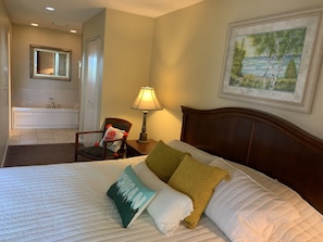Une chambre d’hôtel avec un grand lit, une chaise, une lampe, un tableau et une salle de bain avec une baignoire.