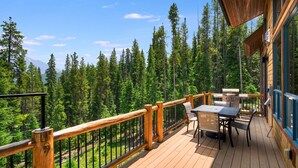 Eine Holzterrasse mit Tisch und Stühlen, umgeben von einem Geländer, mit Blick auf eine bewaldete Landschaft und einen Berg im Hintergrund.
