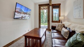 Ein gemütliches Wohnzimmer mit einem Holztisch, einer Ledersofa und einem großen Fernseher, der eine verschneite Landschaft zeigt.