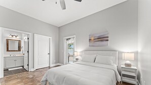 A bedroom with a large bed, a nightstand with a lamp, a bathroom with a mirror and sink, and a view of the outdoors through a window. - Panama City Beach House with Heated Private Pool, Gulf Views, & Steps to Beach! (Panama City Beach)