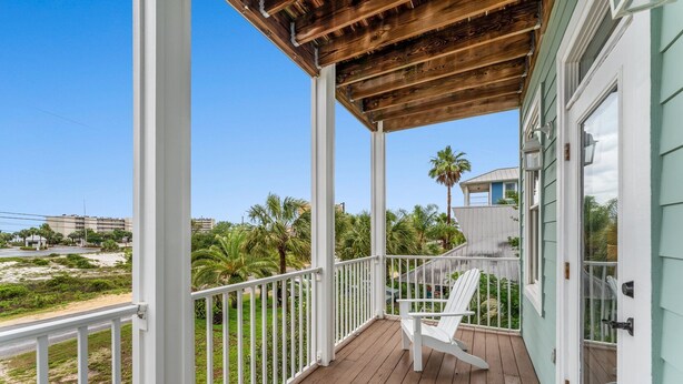 A wooden deck with a white railing, a white chair, and a view of palm trees and buildings in the distance. - Panama City Beach House with Heated Private Pool, Gulf Views, & Steps to Beach! (Panama City Beach)