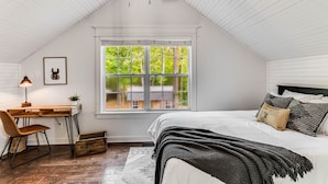 Ein Schlafzimmer mit schrägen Wänden, einem Holztisch, einem Stuhl, einem Bett mit weißer Bettdecke und einem Fenster mit Blick auf ein Haus.