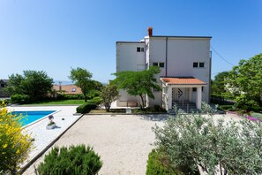 Um edifício moderno de dois andares com piscina, rodeado por jardins e árvores bem cuidados.