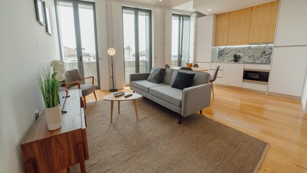 Una sala de estar moderna con un sofá gris, una mesa de centro y un aparador de madera. Se ve un ventanal con vistas a edificios.