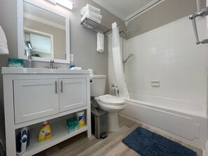 Ein modernes Badezimmer mit Waschbecken, Spiegel und Toilette in Weiß. Eine Dusche mit Glastür und eine blaue Fußmatte sind vorhanden.