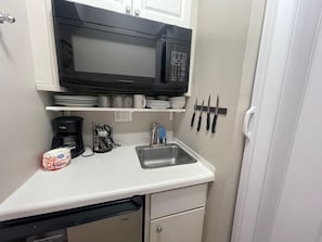 Eine kompakte Küchenzeile mit einer schwarzen Mikrowelle, einer Kaffeemaschine, einem Spülbecken und einem Messerset an der Wand.