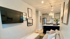 A modern living room with a grey sofa, a coffee table, and a green rug. There is a flat-screen TV mounted on the wall, and a kitchen area is visible in the background. - Pikes Retreat - Mountain Views + Modern Comfort - Hot tub (Colorado Springs)