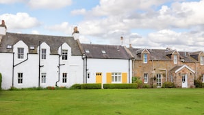 A row of traditional houses with white and stone facades, a grassy lawn, and a partly cloudy sky. - Two bed period cottage in Gullane sleeping up to 4 guests. (Gullane)