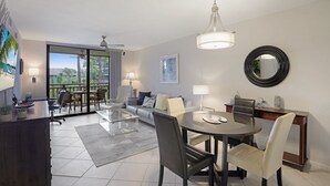 Ein modernes Wohnzimmer mit Glastisch, Sofa und Essbereich mit Stühlen. Ein Balkon mit Blick auf Palmen und Deckenventilator.
