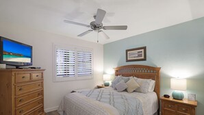 Ein Schlafzimmer mit einem Bett, Holznachttischen, einem Deckenventilator, einem Fernseher und einem Fenster mit Jalousien.