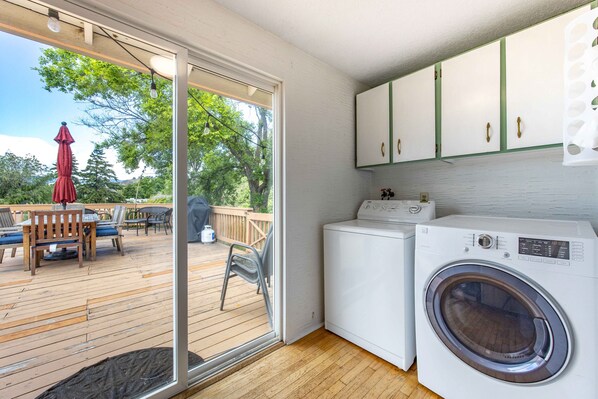 A laundry room with a washing machine and dryer, a sliding glass door to a deck, and a small outdoor seating area with a table and chairs. - Point Lyle - River Views, Pellet Stove, Scenic! (Lyle)