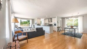 A spacious living area with a wooden floor, a sofa, a dining table with chairs, and a kitchen in the background.