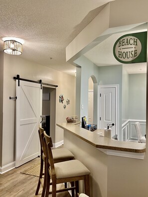 A kitchen with a bar area, a sliding barn door, and a sign that reads "Beach House". - Cozy 3bd/2bh at Barefoot Resort & Golf ⛳️ 🏝 ☀️ 5 min to beach! (North Myrtle Beach)
