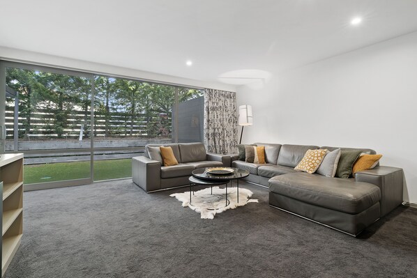 A modern living room with a grey sectional sofa, two armchairs, a glass coffee table, and a large window offering a view of a fenced outdoor area.