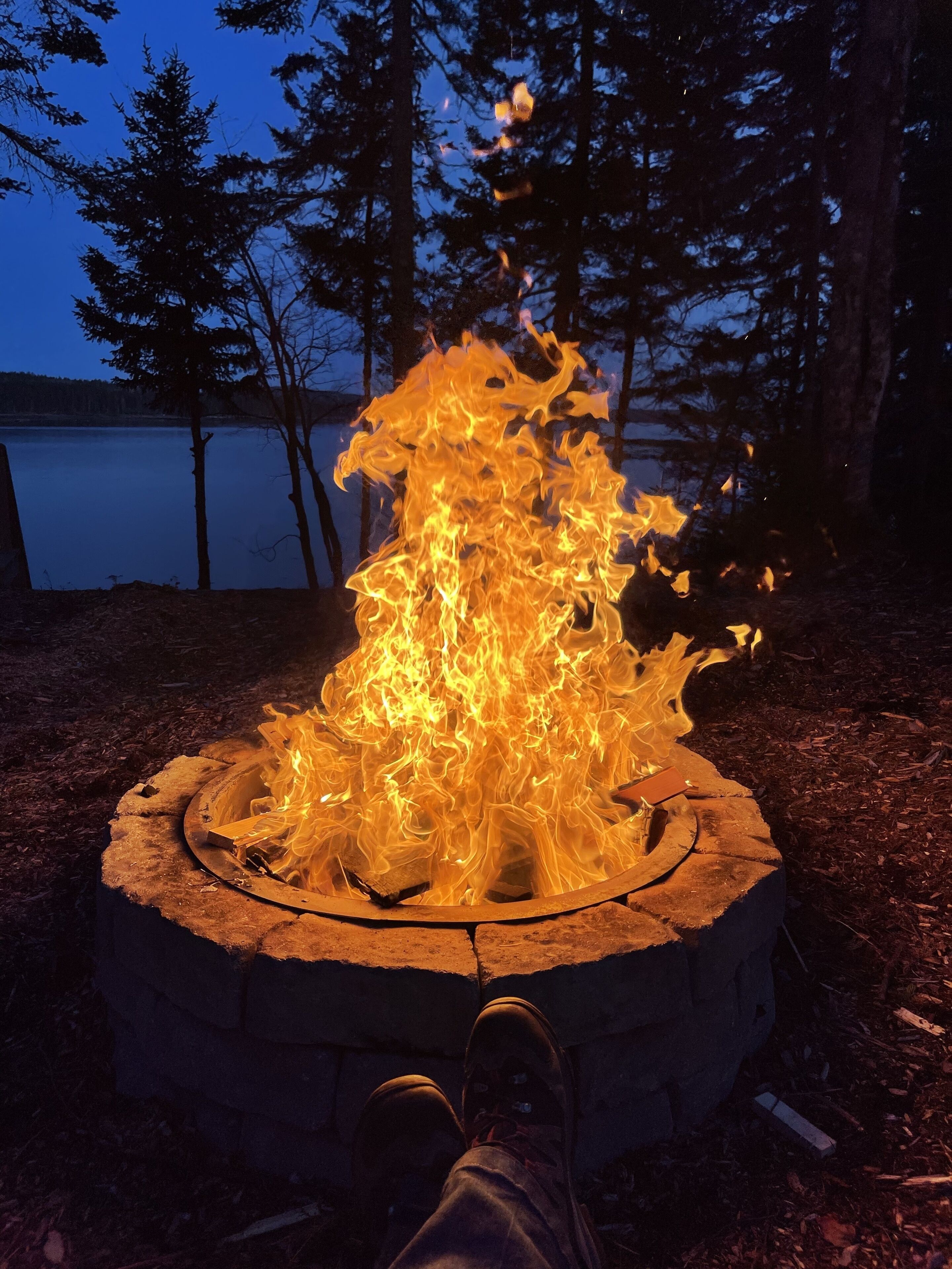 Ein Lagerfeuer in einer runden Steinfürstelle, umgeben von bewaldetem Gebiet mit einem See im Hintergrund.