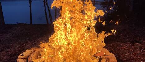 Ein Lagerfeuer in einer runden Steinfürstelle, umgeben von bewaldetem Gebiet mit einem See im Hintergrund.