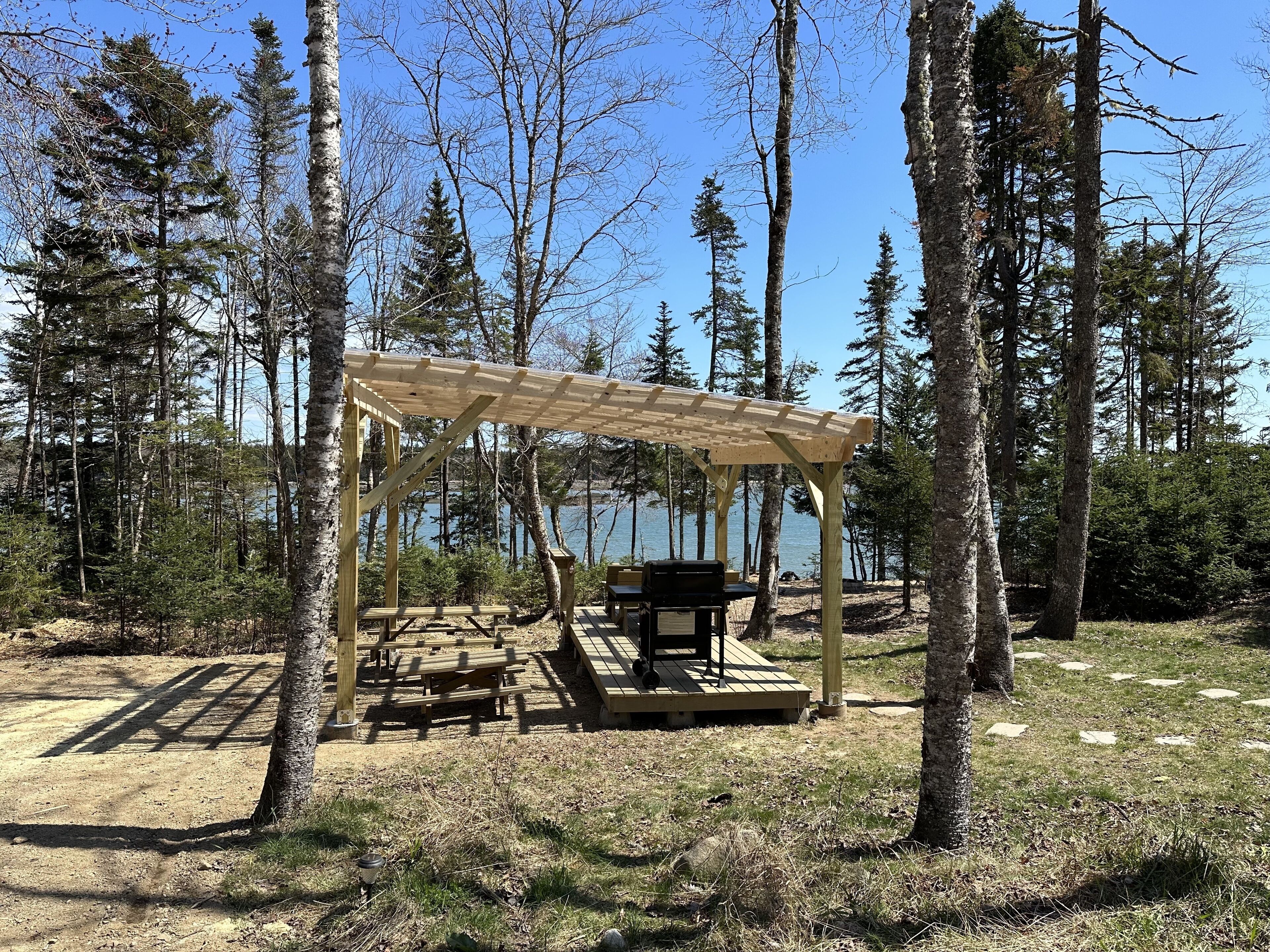 A wooden gazebo with benches and a table, surrounded by trees and a lake.