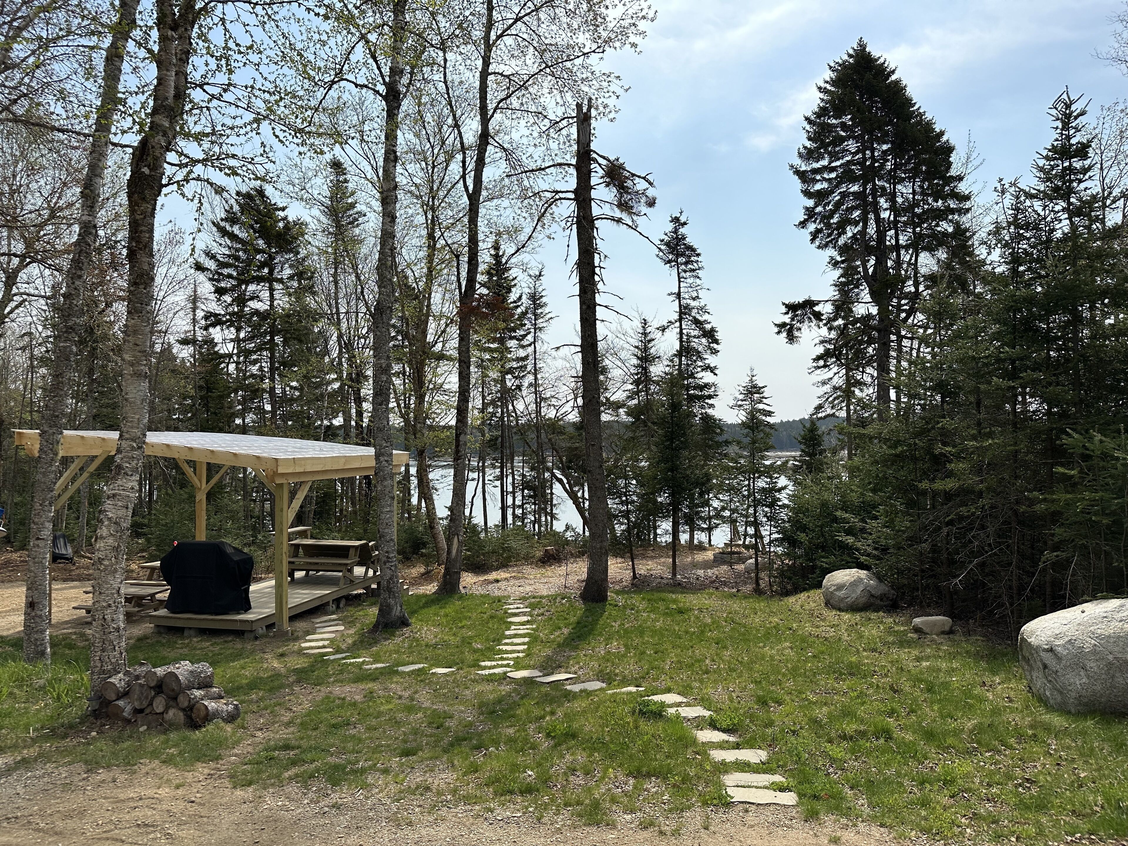 A wooden deck with picnic tables and a barbecue grill, surrounded by a forest and a lake.