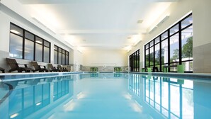 Una piscina interior con agua cristalina y azul, rodeada de amplias ventanas y sillones de descanso.