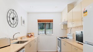A modern kitchen with a sink, stove, and microwave. There is a window with blinds, a clock on the wall, and a cabinet with a sticker. - TOWN CENTRE LOCATION WITH STUNNING VIEW - UNIT 1 (Queenstown)