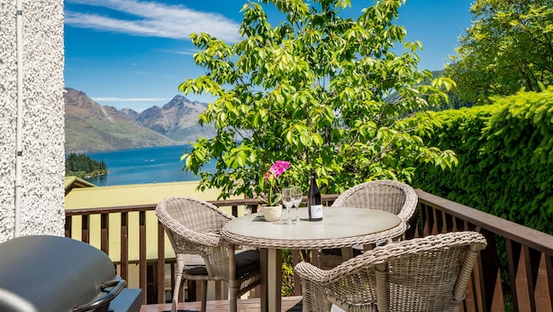 A balcony with wicker furniture, a table with a bottle and glass, and a view of a lake and mountains. - TOWN CENTRE LOCATION WITH STUNNING VIEW - UNIT 1 (Queenstown)