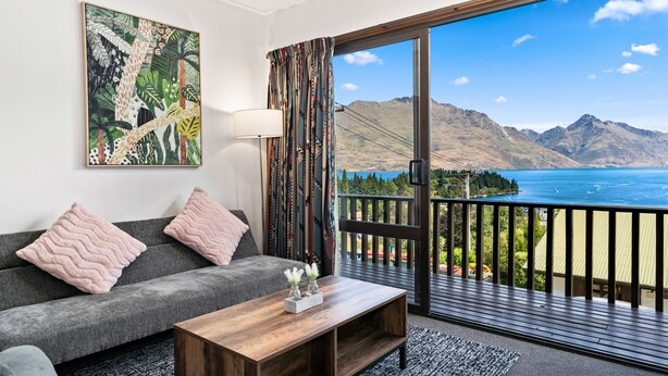 A room with a grey sofa, two pink pillows, a wooden coffee table with a vase, a painting of tropical foliage, and a balcony with a mountain view. - TOWN CENTRE LOCATION WITH STUNNING VIEW - UNIT 8 (Queenstown)