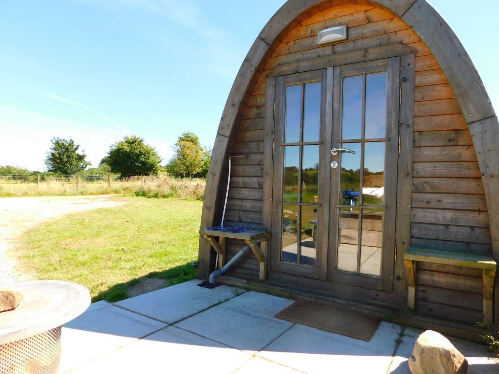 Una cabaña de madera con una puerta y ventanas de arco grande, una pequeña banca en el exterior y una zona de patio empedrada.