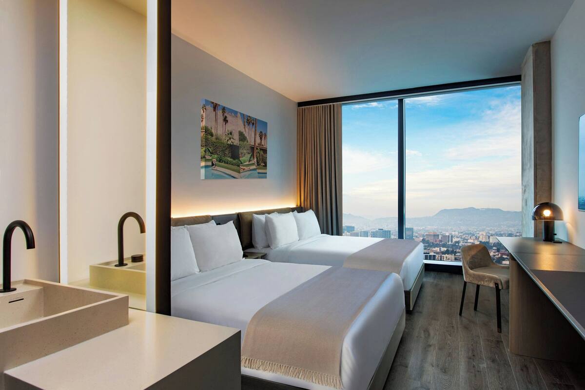 A modern hotel room with two beds, a desk, a chair, a large window with a city view, and a painting on the wall.
