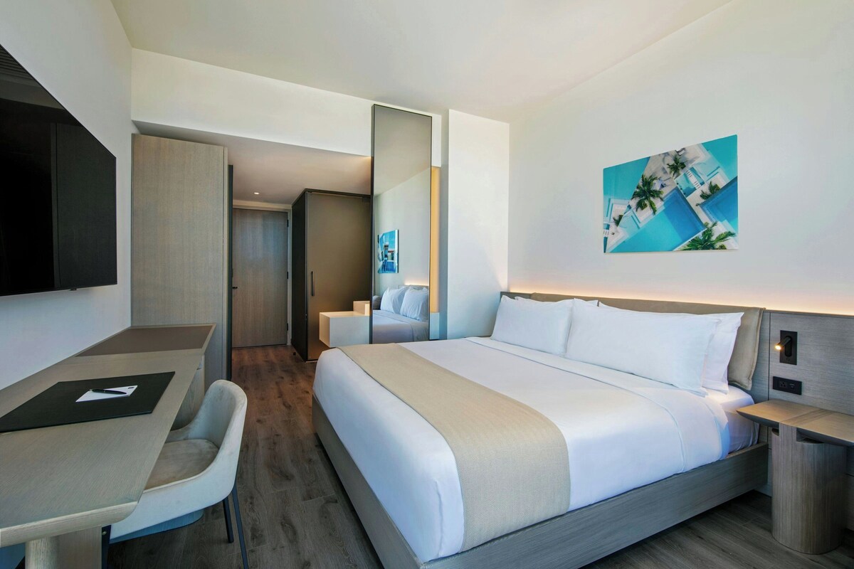 A modern hotel room with a large bed, a desk, and a chair. There is a TV mounted on the wall and a painting above the bed.