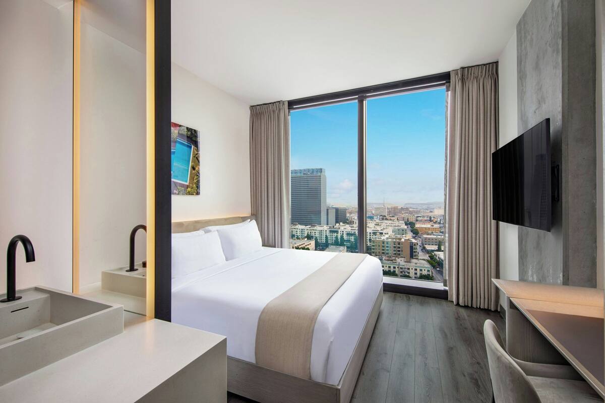 A hotel room with a large bed, a TV, a desk, and a view of the city through large windows.