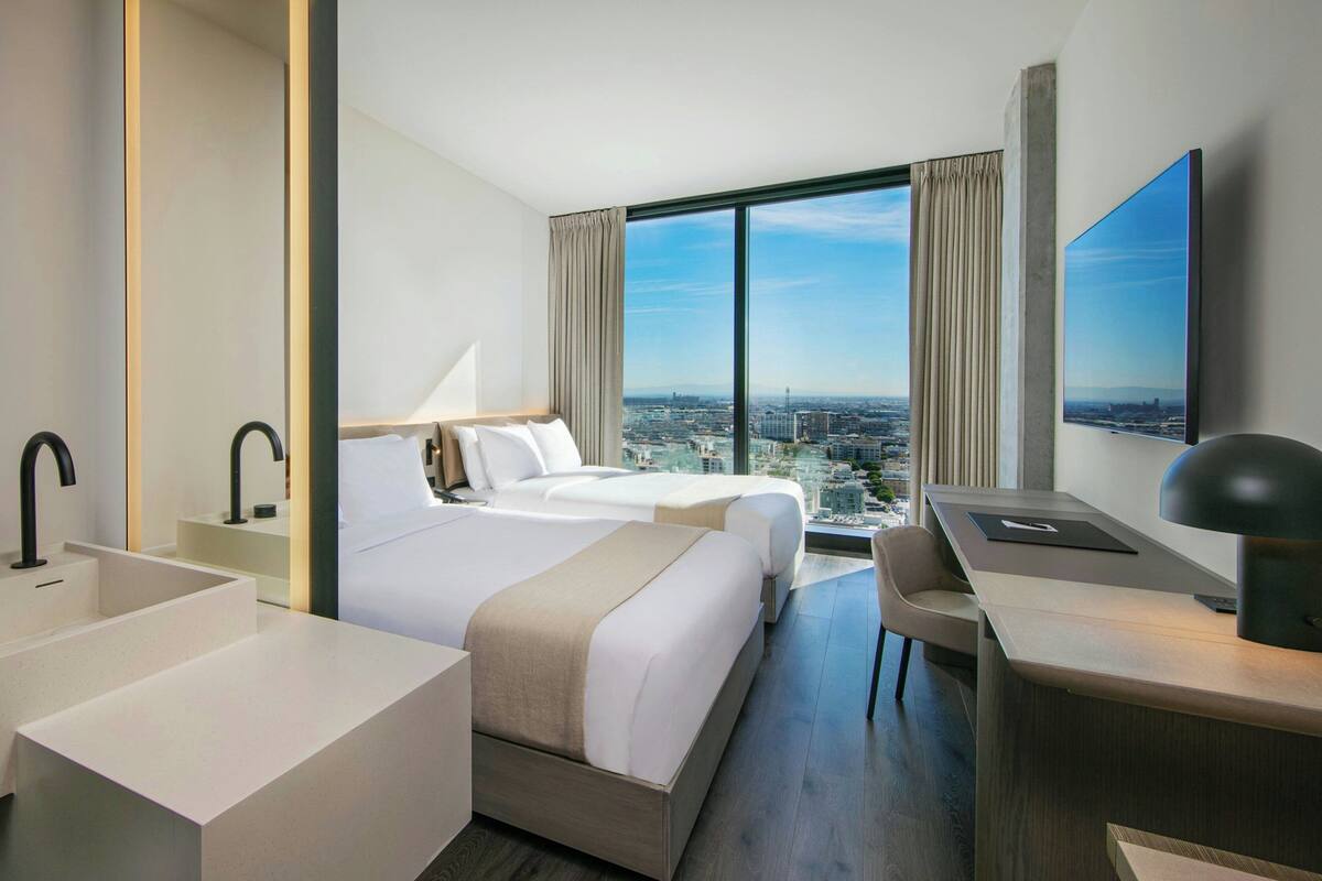 A hotel room with two beds, a desk, a large window with a city view, a bathtub, and a large painting on the wall.
