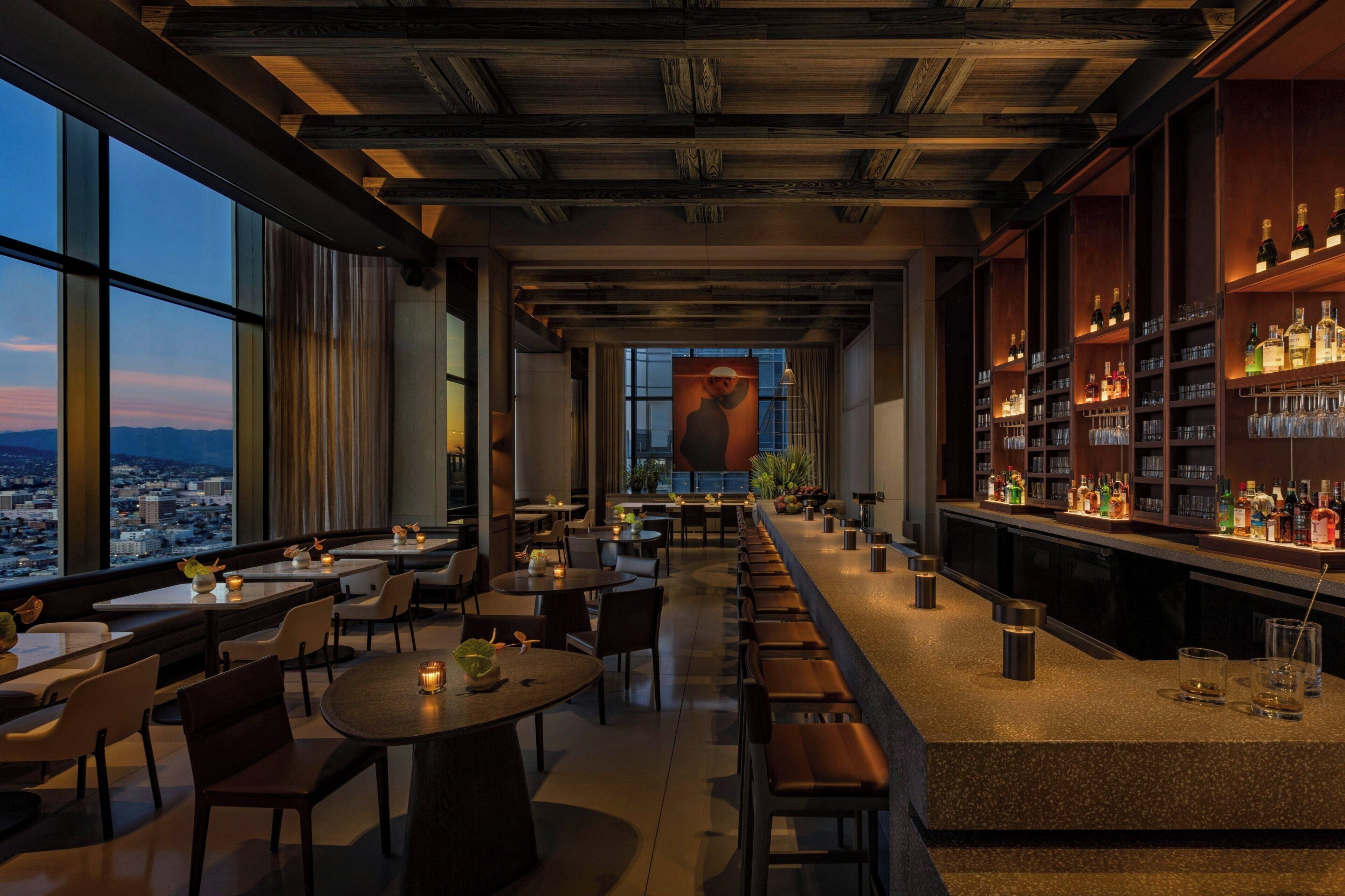 A modern bar area with a long counter, seating, and a view of the cityscape.