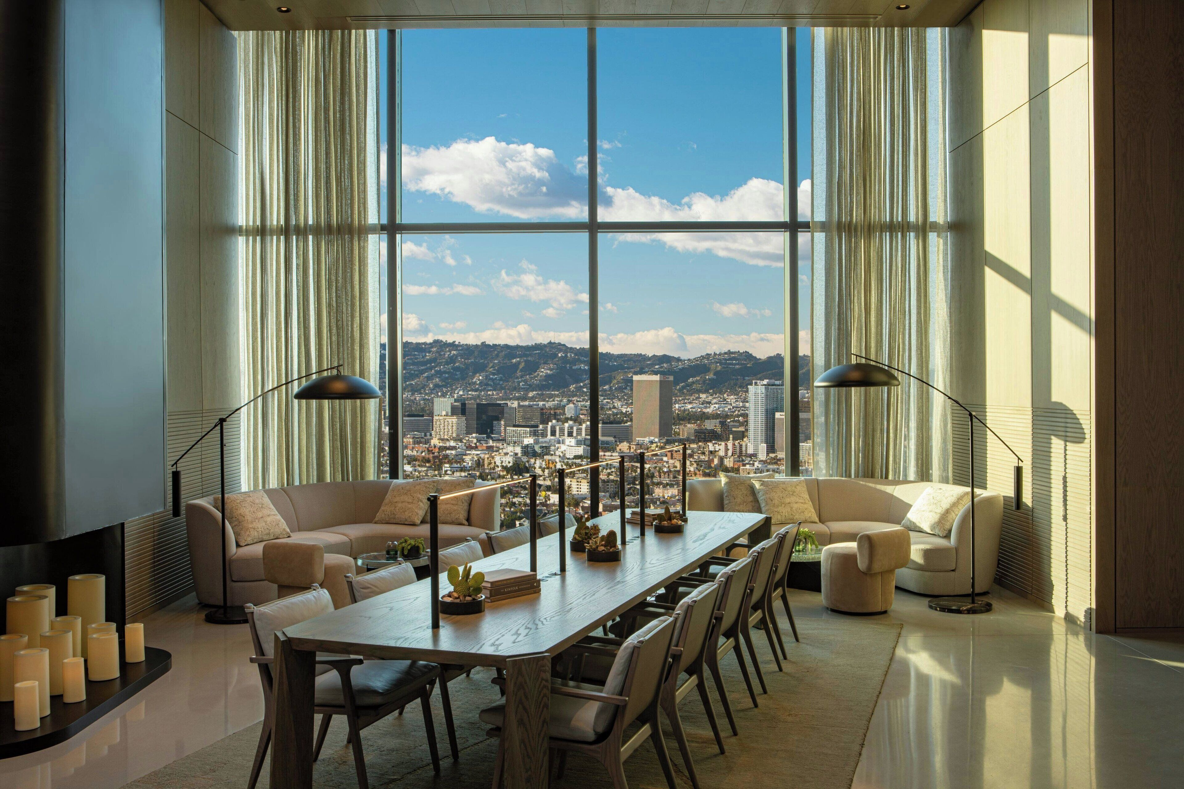 A spacious living room with a large window offering a city view, a long dining table, and modern furniture.