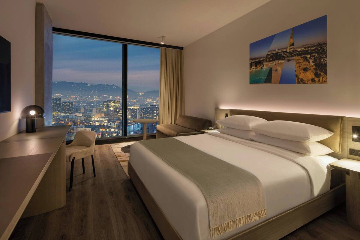 A modern hotel room with a large bed, a desk, and a view of a cityscape through a large window.