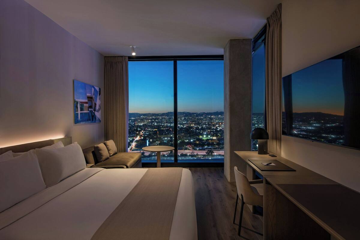 A hotel room with a large bed, a desk, a chair, a small table, and a large window offering a city view.
