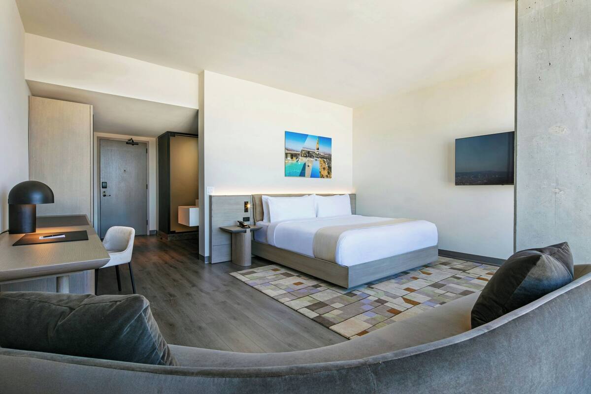A modern hotel room with a bed, a desk, a chair, a sofa, and a television.