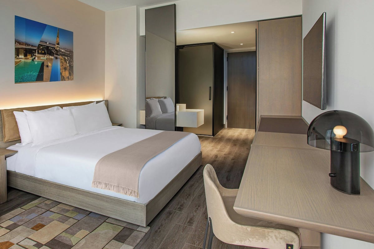A modern hotel room with a large bed, a desk, and a chair. There is a painting on the wall and a door leading to another room.