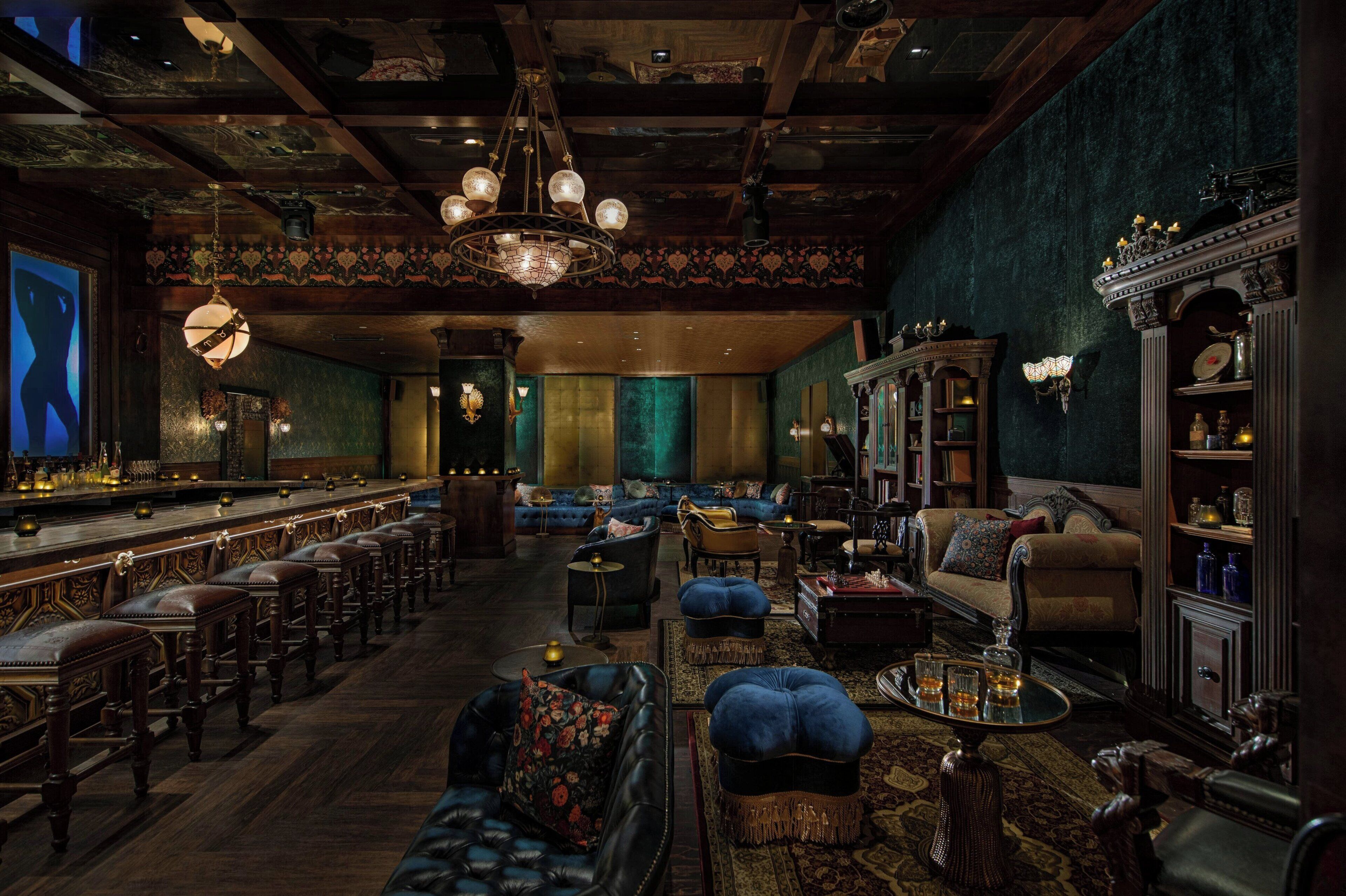 Un espace bar avec des tabourets de bar en bois, un long comptoir et un lustre. Des canapés et des fauteuils sont disposés dans l’espace détente.