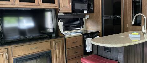 A compact kitchen with wooden cabinets, a microwave, a stove, a sink, and a flat-screen TV.
