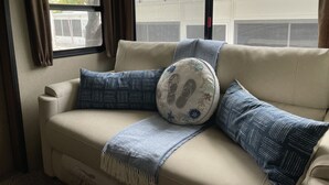 Ein beiges Sofa mit gemusterten Kissen und einer Decke, platziert neben einem Fenster mit Vorhängen.