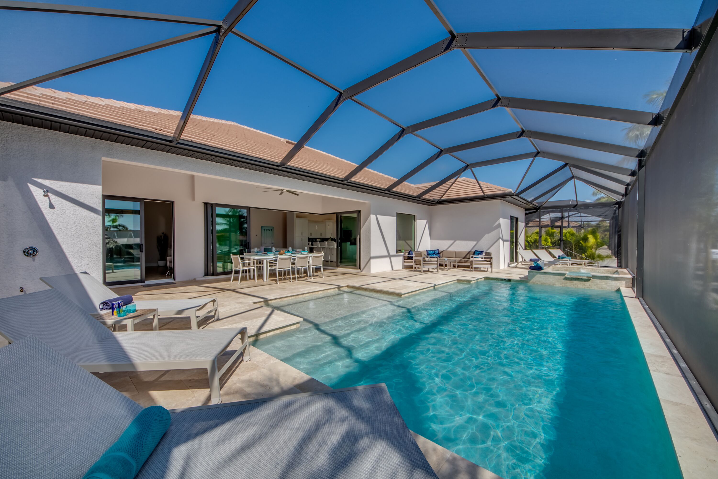 Un espace piscine extérieur moderne doté d’un toit en verre, de chaises longues et d’un coin repas.