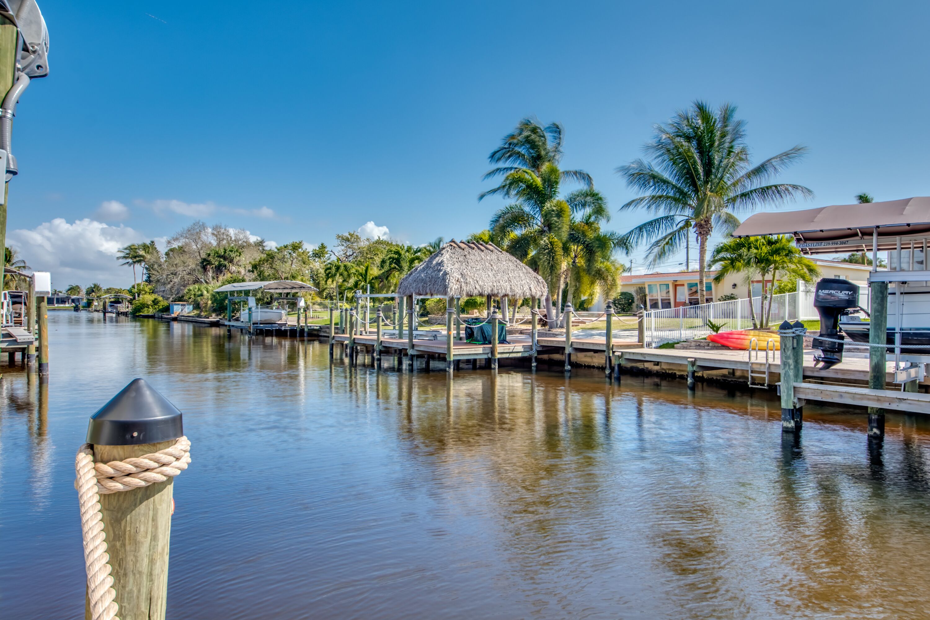 A waterfront dock with a thatched-roof structure, palm trees, and a boat.