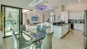 Una cocina moderna con armarios blancos, una isla central y un comedor con mesas y sillas de vidrio.