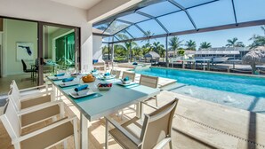 Zona de comedor junto a la piscina con un porche de vidrio, una piscina y vistas a palmeras y una playa.