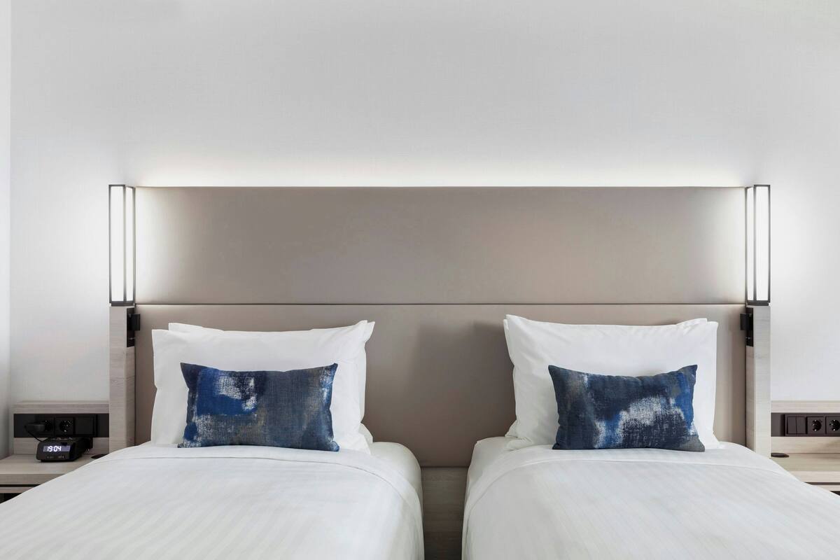A hotel room with two beds, each with white bedding and a blue abstract pillow. There is a nightstand with a clock and a lamp on each side of the beds.