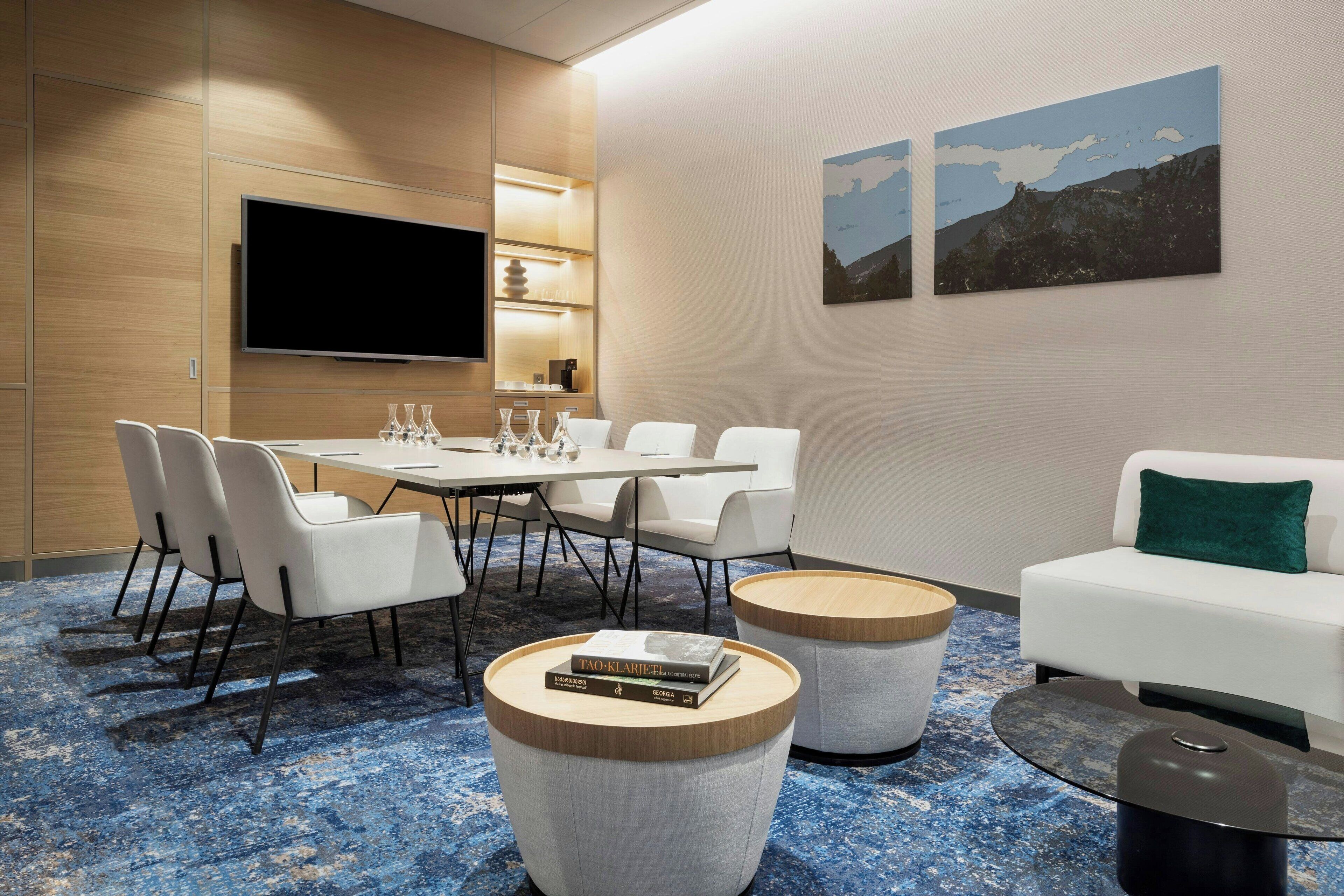 Habitación de hotel moderna con zona de comedor, televisor de pantalla plana, dos cuadros y un sofá blanco.
