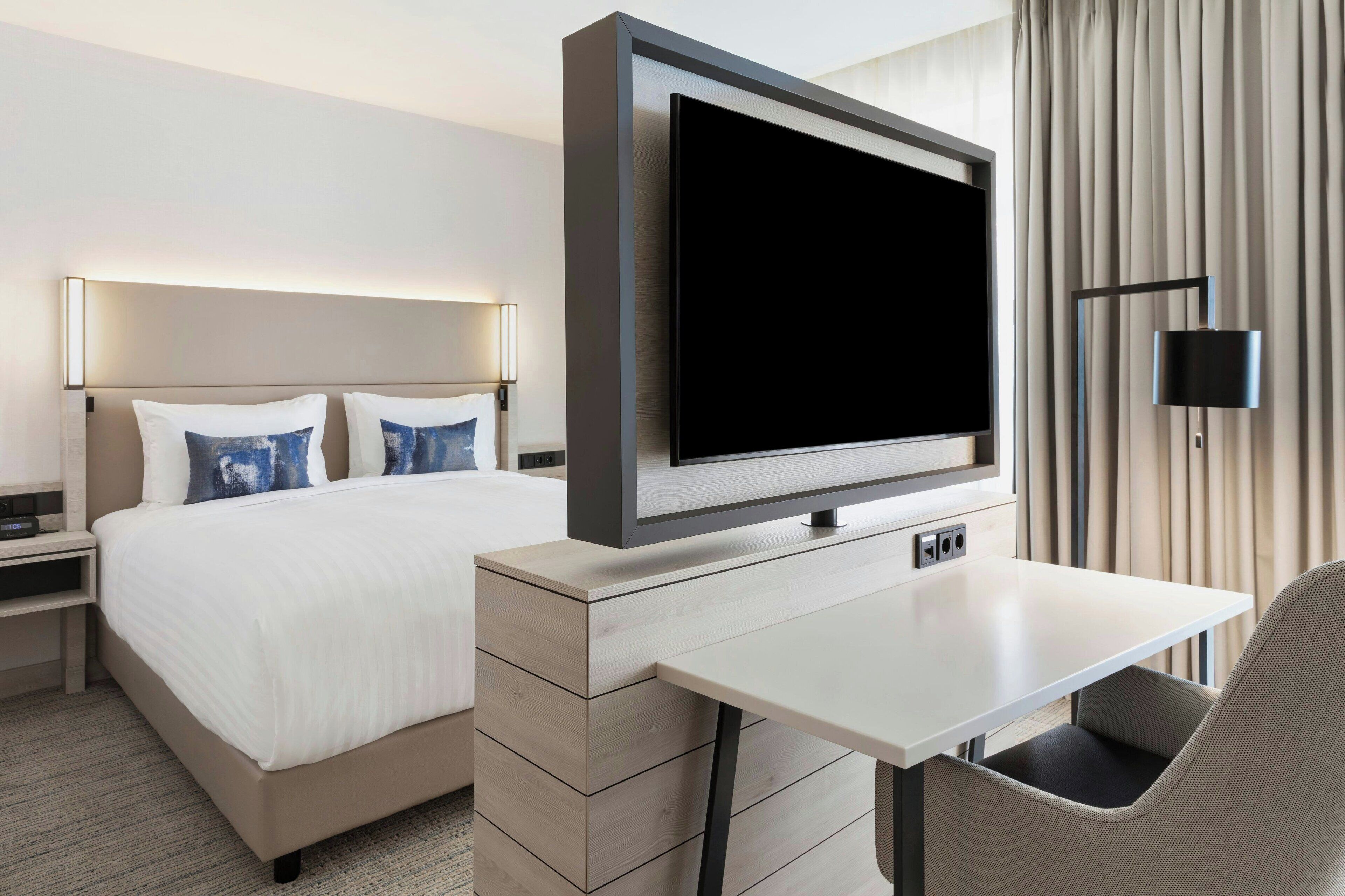 Una habitación de hotel moderna con una cama grande, un televisor de pantalla plana en la pared, un escritorio con una silla y una lámpara.